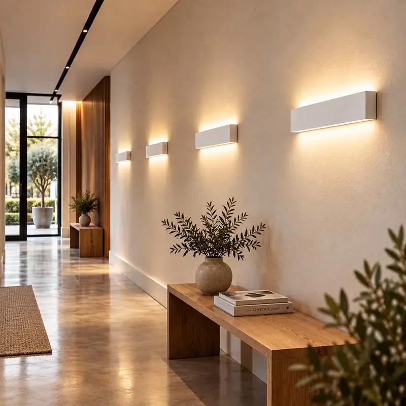 Modern rectangular wall sconces mounted on a beige wall in a minimalist hallway, casting a warm glow. A wooden bench with a vase of greenery and books sits below.