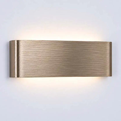 A modern rectangular wall sconce with a brushed brass finish, emitting a warm glow upwards and downwards against a plain light gray wall.