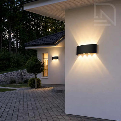 Modern black outdoor wall sconces mounted on a white stucco house exterior at dusk, casting warm light onto the wall and driveway.