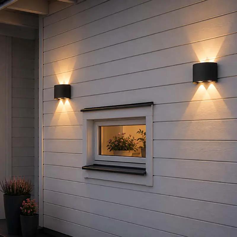 Two black cylindrical outdoor wall sconces mounted on a white horizontal siding house wall, casting warm light downward and upward around a small window.
