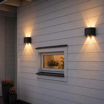 Two black cylindrical outdoor wall sconces mounted on a white horizontal siding house wall, casting warm light downward and upward around a small window.