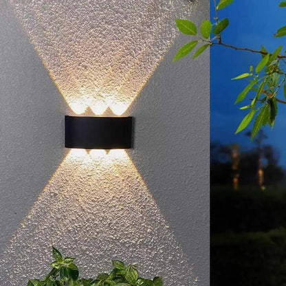 A modern black rectangular outdoor wall sconce mounted on a textured stucco wall, casting a warm light upward and downward in a V-shape. A split-screen view on the right shows the same fixture in use at dusk against a dark blue sky.