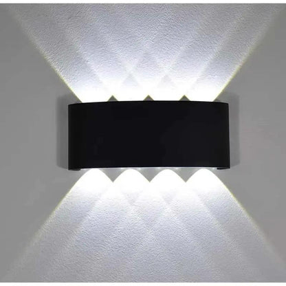 A modern black wall-mounted light fixture with three upward-facing and three downward-facing illuminated beams, shown against a textured white wall.