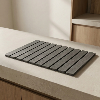 A rectangular dark gray slatted mat or trivet placed on a light beige countertop in a kitchen setting with wood cabinetry visible in the background.