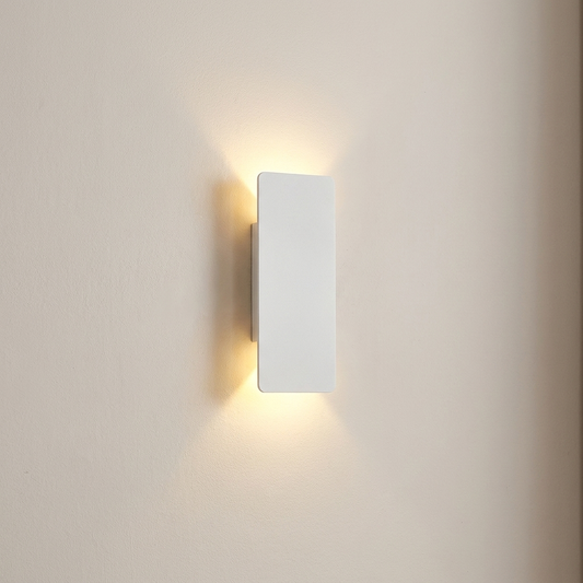 A minimalist rectangular wall sconce light fixture in matte white, mounted on a warm beige wall and casting a soft warm yellow glow downward and outward.