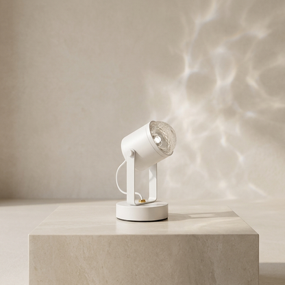 A modern white adjustable spotlight lamp with a clear glass shade, resting on a light beige stone pedestal. The lamp is turned on, casting a soft, rippled water-like shadow pattern on the beige wall behind it.